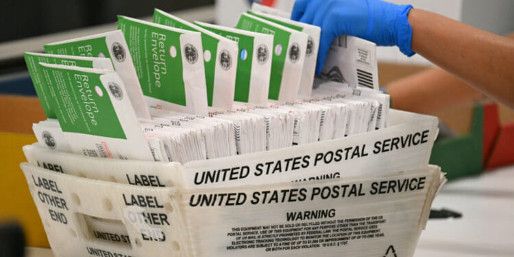 A gloved hand handles a stack of mail-in ballots in green and white envelopes inside a United States Postal Service tray.