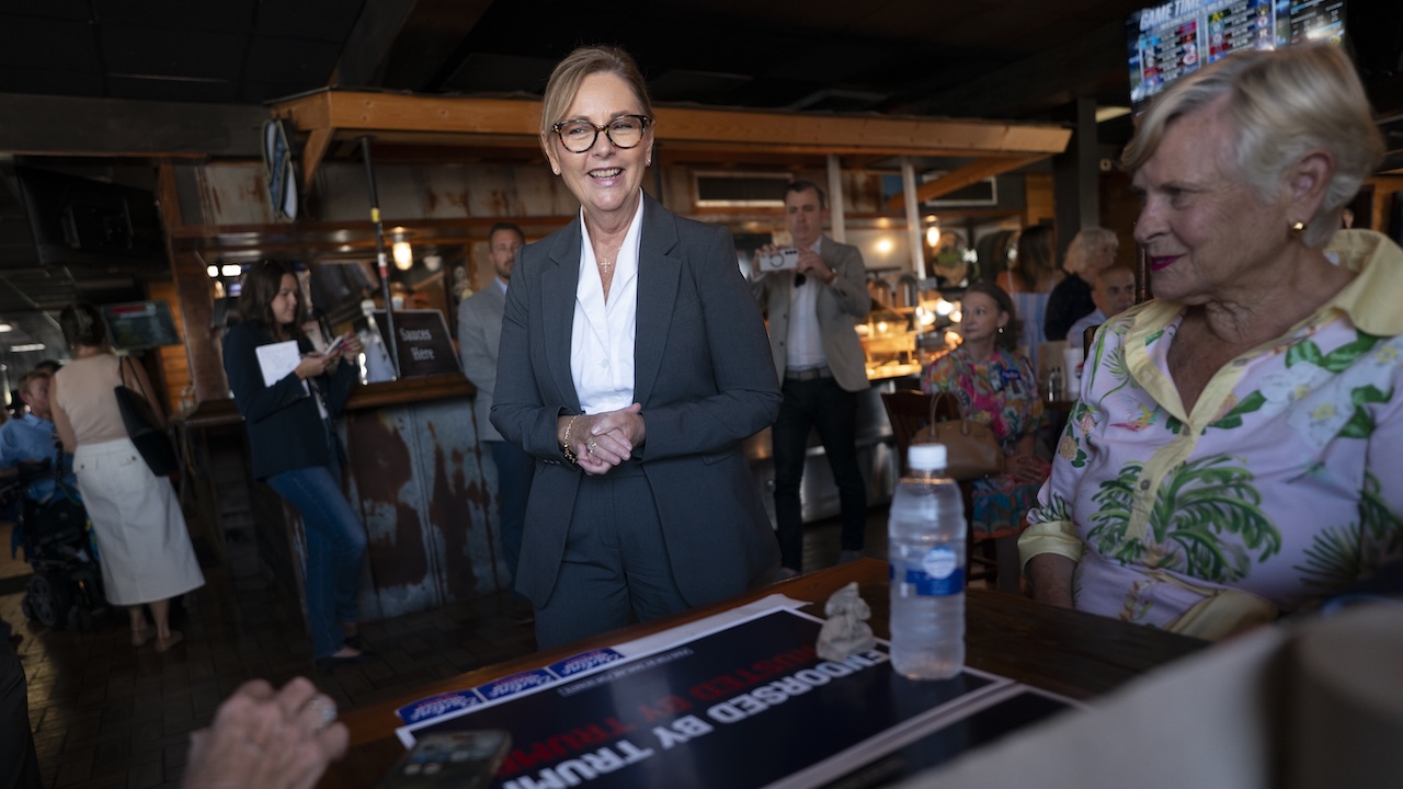 A woman in a suit smiles while standing near a table with a campaign sign and water bottle. People sit and mingle in a casual indoor setting with warm lighting.