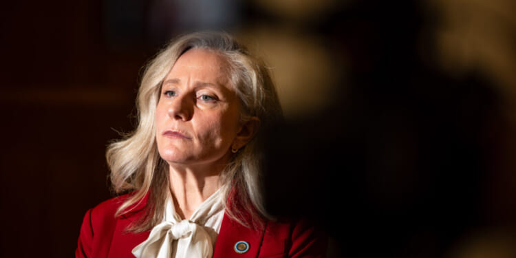 Abigail Spanberger with long gray hair, who wears a red blazer and white blouse, looks slightly to the side. The background is dimly lit with a dark, shadowy area on the right.
