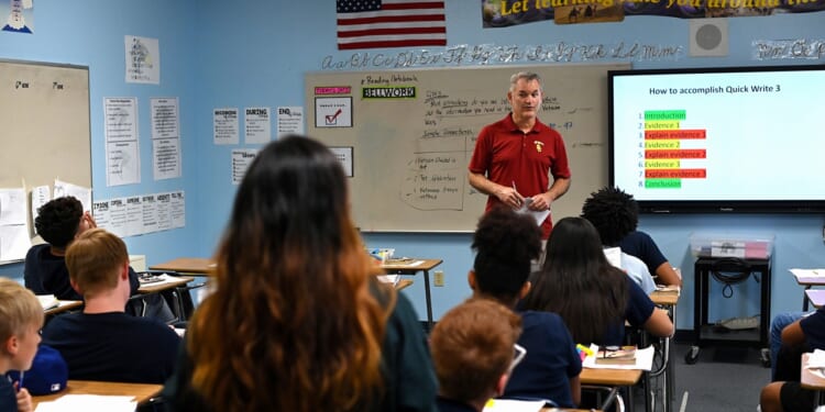 A teacher stands at the front of a classroom, speaking to students seated at desks. A large screen displays instructions, and educational posters are visible on the walls. An American flag hangs in the background.