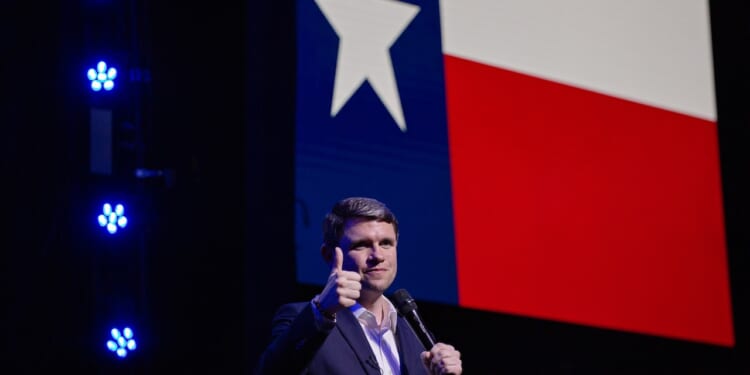 A man in a suit gives a thumbs up while holding a microphone, standing in front of a large Texas flag projected on a screen. Blue stage lights are visible on the left.