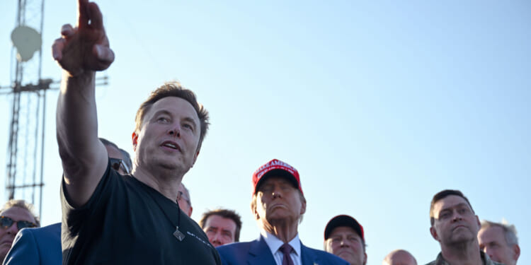Elon Musk in a black shirt gestures while speaking to Donald Trump and a group of people outdoors.