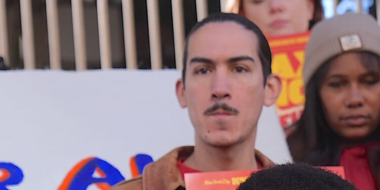 A person with a mustache stands in front of a white protest sign, holding a red pamphlet. Others are visible in the background, including a woman in a beanie. The mood appears serious and focused.
