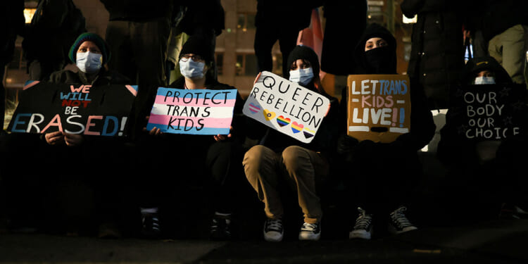 The transgender movement's arguments in court have changed from aggressive promotion to defense. Pictured: Five people sitting, holding protest signs with messages supporting trans and queer rights, such as Protect Trans Kids and Let Trans Kids Live. All are wearing masks, and their faces are partially obscured by shadows.