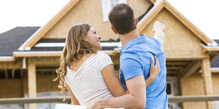 Homeownership concept. Pictured: a couple stands in front of a partially built house, embracing. The man points toward the home while the woman holds rolled blueprints, both looking at the construction with anticipation.