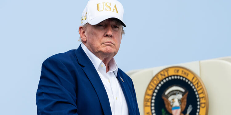 A man in a blue suit and white USA hat stands in front of a partial view of the presidential seal.
