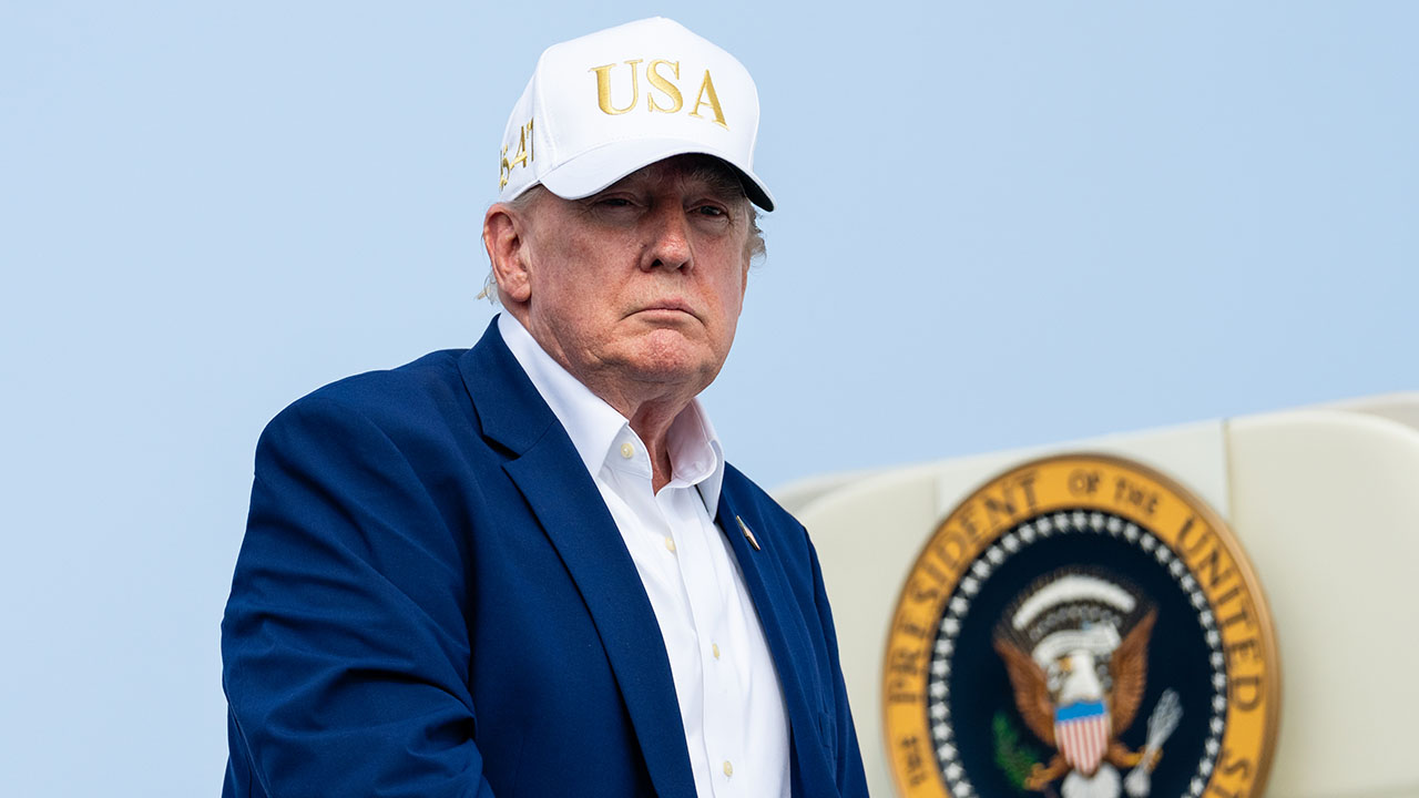 A man in a blue suit and white USA hat stands in front of a partial view of the presidential seal.