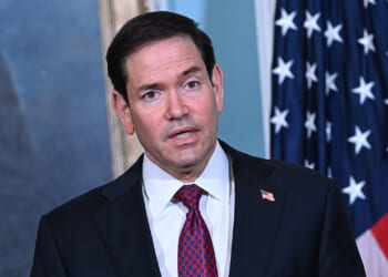Marco Rubio in a suit and red patterned tie speaks in front of a U.S. flag and a painting, with an American flag pin on his lapel.