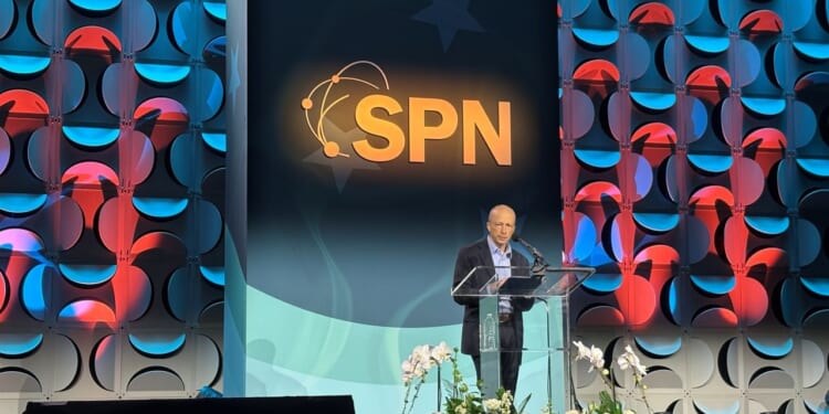 Speaker Yuval Levin stands at a podium on a decorated stage with the letters SPN prominently displayed behind him. Flowers are arranged in front of the podium, and the background features a geometric pattern with blue and red lights.