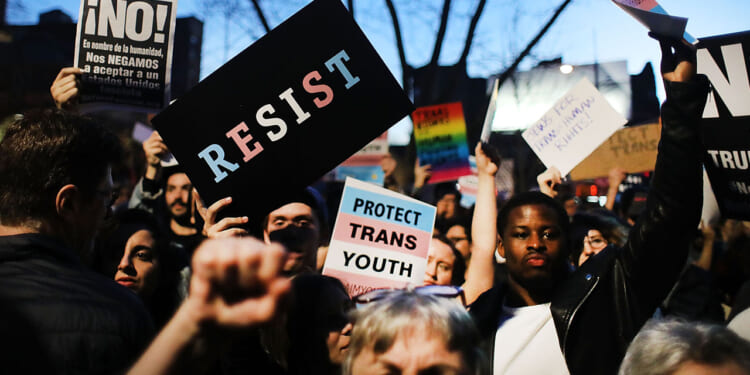 The World Professional Association for Transgender Health just made a key admission. WPATH said its Standards of Care are opinion, not objective fact claims. Pictured: a crowd of protesters holds signs, including RESIST, PROTECT TRANS YOUTH, and Spanish-language messages. People have raised fists, and a trees branches are visible in the background at dusk.
