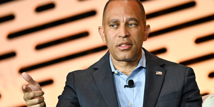 Hakeem Jeffries, wearing a suit, gestures with his left hand while speaking, seated in front of a blurred background with horizontal black and orange patterns.