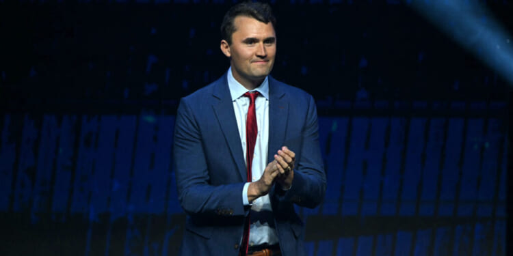 Charlie Kirk, dressed in a blue suit and red tie, stands on stage, clapping his hands, with a dark blue background and a spotlight shining from the upper right corner.