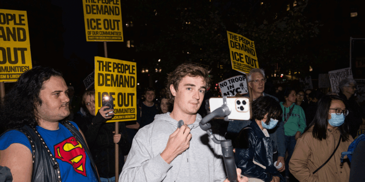 Nick Shirley speaks into a microphone connected to a phone at a nighttime protest. People around him hold yellow signs reading THE PEOPLE DEMAND ICE OUT OF OUR COMMUNITIES. The crowd includes diverse ages and attire.