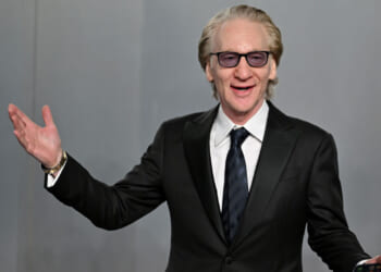 A man in a black suit, white shirt, and dark tie smiles and gestures with one hand raised. He has light hair, wears glasses, and stands against a plain gray background.