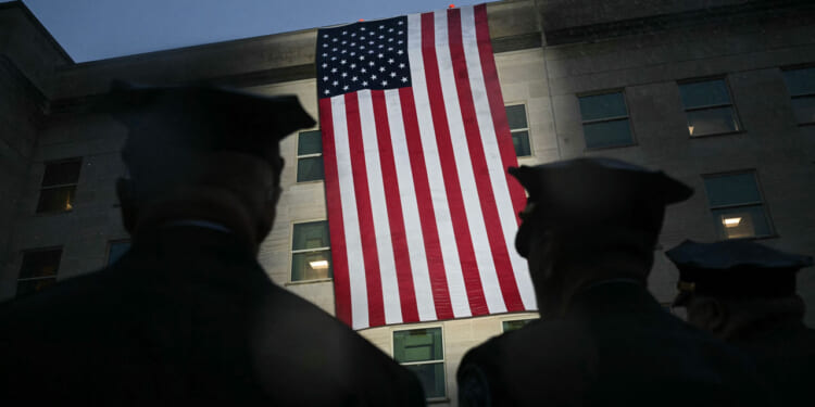 Silhouetted figures in uniform stand facing a large American flag hanging on the side of a building at dusk.