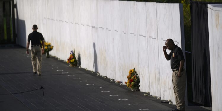 Two security guards stand by a white marble memorial wall with engraved names and flowers placed at its base on a sunny day.