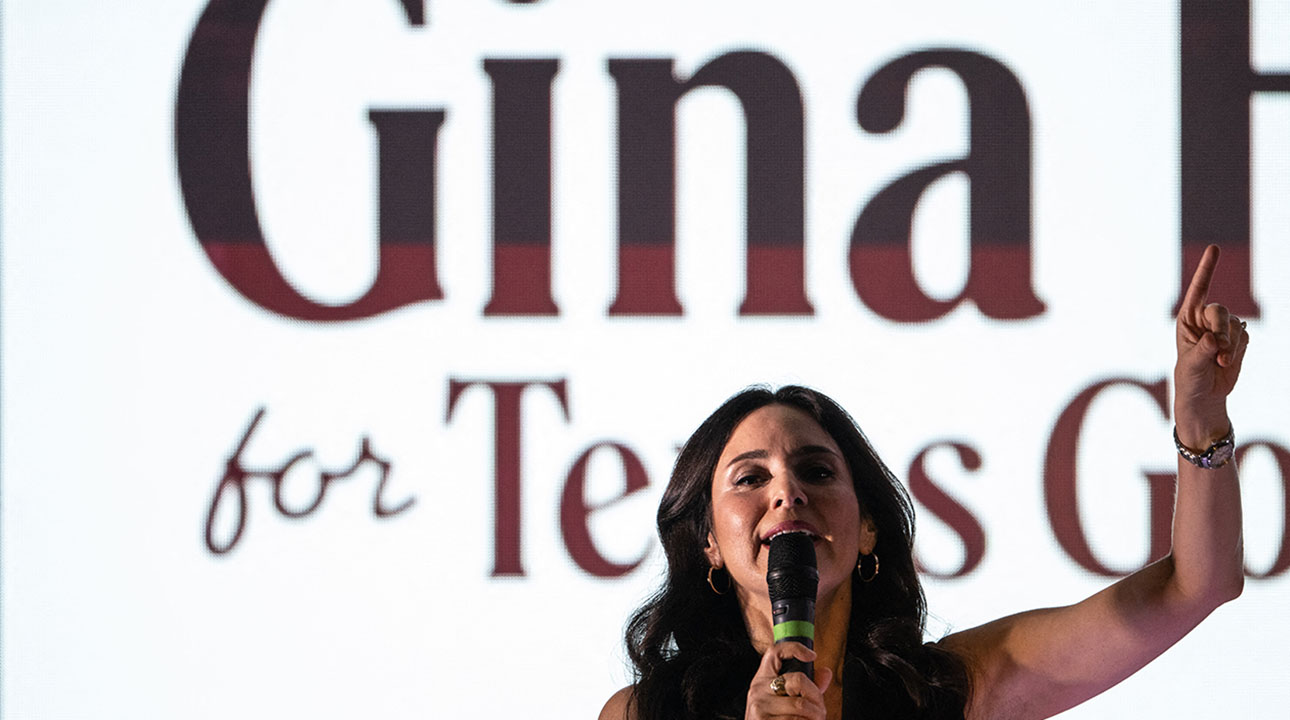 A woman with dark hair speaks into a microphone, raising one finger, in front of a large screen displaying partially visible text that reads Gina and for Texas..