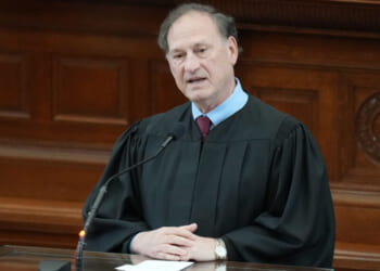 A judge in a black robe speaks at a podium in a courtroom, with a microphone in front of him and wooden paneling in the background.