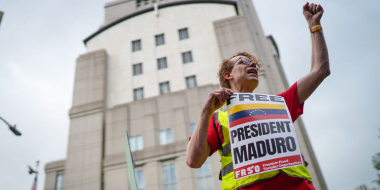 A person wearing glasses and a Free President Maduro sign raises a fist during a protest in front of a tall building.