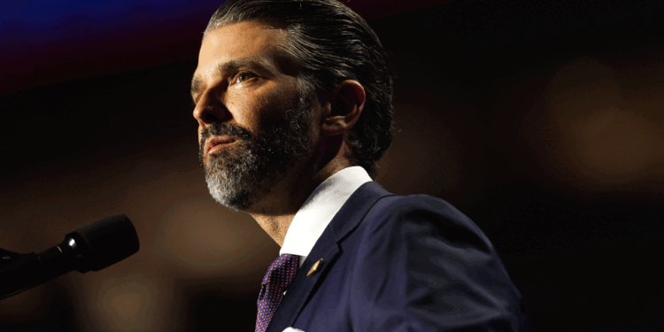 Don Trump Jr. in a suit and tie with slicked-back hair and a beard stands at a microphone, looking serious, with a dark blurred background.