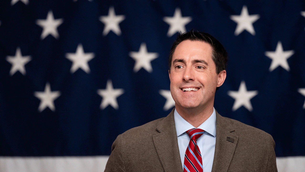 A man in a suit and red striped tie smiles in front of a backdrop with white stars on a blue background, resembling part of the American flag.