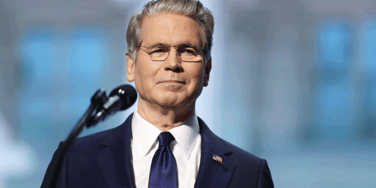 Scott Bessent in a navy suit and tie stands in front of a microphone, looking forward with a neutral expression. He has gray hair, glasses, and an American flag pin on his lapel. The background is blurred.