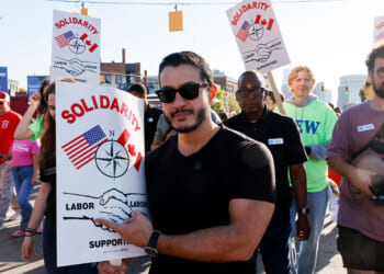 A group of people march on a street holding Solidarity signs with US and Canadian flags, a compass, and shaking hands, supporting labor unions and advocating for fair election practices. Abdul El-Sayed in sunglasses holds a sign in the front.