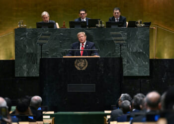 A man in a suit speaks at a podium with the United Nations emblem, addressing an audience. Four officials sit behind him on a raised platform in a large, formal assembly hall.