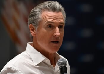 A man with gray hair wearing a white shirt speaks into a microphone. An out-of-focus American flag is visible in the background.