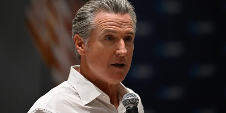 A man with gray hair wearing a white shirt speaks into a microphone. An out-of-focus American flag is visible in the background.
