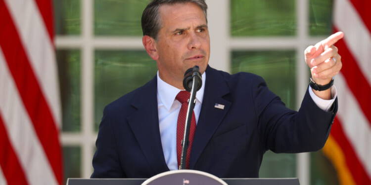A man in a dark suit and red tie speaks at a podium outdoors, gesturing with his right hand as he addresses concerns about election fraud. American flags and green foliage are in the background.