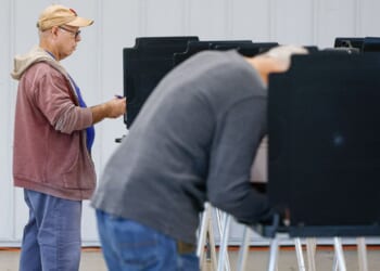 Two people are standing at voting booths, filling out ballots. The scene is indoors, with a white wall in the background. One person is in focus, while the other is blurred in the foreground.