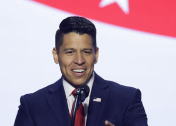A man in a dark suit and red tie speaks at a podium with a microphone, against a backdrop featuring part of a red stripe and a white star, representing the Texas 35th Congressional District.