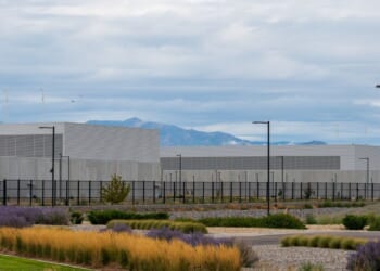 A large, modern industrial data center building with gray walls and flat roofs. It sits behind a black fence, surrounded by landscaping with grasses and shrubs, with mountains and cloudy skies in the background.