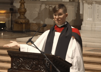 Rev. Flouish Klink claims Jesus is trans. Pictured: a person in religious robes stands at a carved wooden pulpit, speaking into a microphone, with one hand gesturing outward inside a large, ornate church.