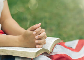 A person sits outdoors with hands clasped in prayer over an open book, draped with an American flag on their lap, with green grass in the background.