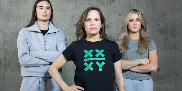 Three women stand confidently against a gray concrete wall. The center woman wears a black XX XY Athletics shirt; the other two wear light-gray athletic outfits with arms crossed. All look directly at the camera.