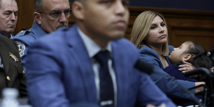 A woman holds a child while seated beside men in suits and uniforms at a formal hearing or meeting, all appear serious and attentive.