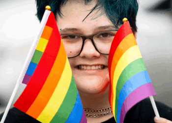 A person with short blue hair and glasses smiles while holding two small rainbow pride flags, one in each hand, partially covering their face.
