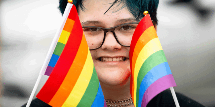 A person with short blue hair and glasses smiles while holding two small rainbow pride flags, one in each hand, partially covering their face.