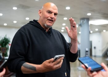 A bald man in a black hoodie gestures with one hand while holding a phone, surrounded by people holding smartphones in an indoor setting.