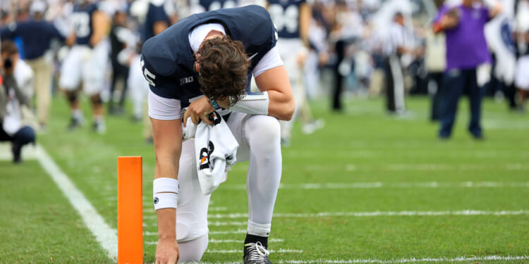 Football player in white pants and a dark jersey kneels on one knee near the end zone, head bowed in an expression of faith and hand touching the ground, with a towel in hand. Crowd and teammates are blurred in the background.