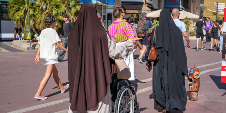 Women in hijabs walk outdoors, one guiding a person in a wheelchair. Other people in casual clothes walk nearby under bright sunlight in an urban setting with shops and palm trees visible.