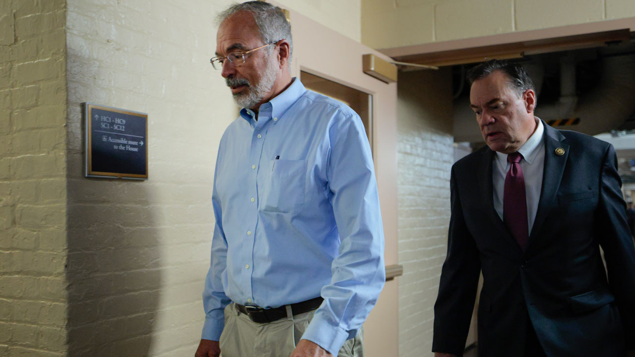 Two men walk through a hallway; one wears a light blue shirt and khaki pants, the other a dark suit and tie. The hallway has beige brick walls and a sign mounted on the left wall.