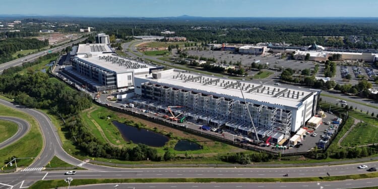 Aerial view of a large industrial data center under construction, surrounded by roads, parking lots, trees, and commercial buildings, with a clear blue sky overhead.