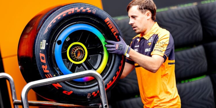 A mechanic in an orange and navy uniform carries a large Formula 1 Pirelli tire with colorful markings, likely for a racing event.