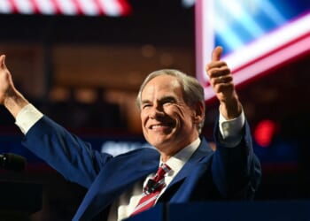 A man in a suit and striped tie smiles and gives two thumbs up while standing at a podium, with bright lights and blurred flags in the background.