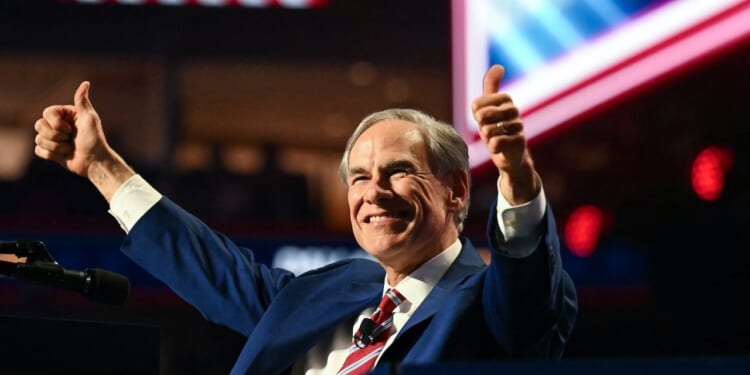A man in a suit and striped tie smiles and gives two thumbs up while standing at a podium, with bright lights and blurred flags in the background.