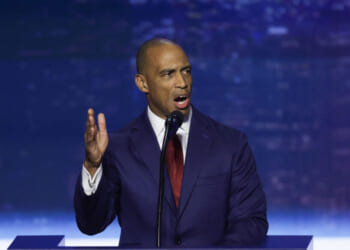 Scott Turner in a navy suit and red tie speaks passionately at a podium, gesturing with his left hand, against a blurred blue and purple background.