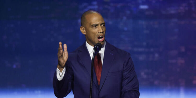 Scott Turner in a navy suit and red tie speaks passionately at a podium, gesturing with his left hand, against a blurred blue and purple background.